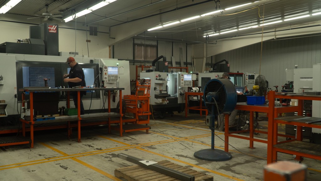 Wide-angle photo of a busy machine shop showing a worker operating a machine amidst various industrial machinery. The scene captures a well-organized workshop with advanced equipment and tools, highlighting a productive environment and skilled craftsmanship.
